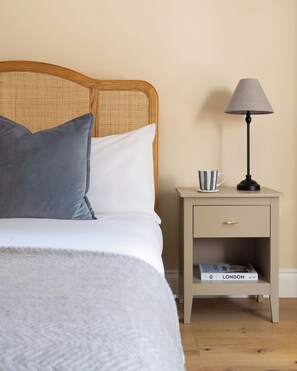 Una cama bien tendida con una manta gris y almohadas, una mesita de noche con una lámpara y una taza a rayas, y un libro titulado 'LONDON' en la estantería.