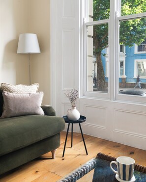 A living room with a green sofa, a white lamp, a small table with a vase, and a view of a blue building through a window. - Notting Hill Grove Apartments (London)
