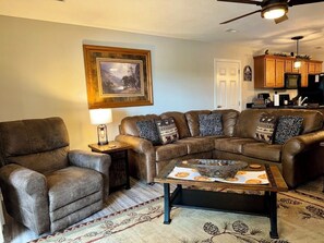 A living room with a brown leather sofa, a brown leather armchair, a coffee table with a placemat, a framed landscape painting, and a kitchen area with wooden cabinets and a microwave. - 3BR lakefront condo with pool, balcony, and resort amenities (Hollister)