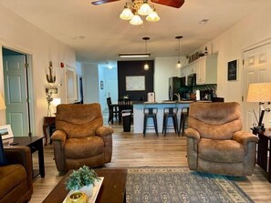 A living room with two armchairs, a coffee table, and a rug. There is a kitchen in the background with a bar and stools. - Lakefront 3BR condo at Table Rock Lake with pool, patio, and Wi-Fi. (Hollister)
