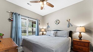 Quarto com cama, duas mesas de cabeceira, um ventilador de teto, janela com cortinas e parede com um círculo de balsa e objetos listrados.
