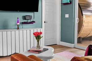 Ein modernes Wohnzimmer mit einer weißen Heizung, einem flachbildfernseher an der Wand, einer pinken Lavalampe, einer Vase mit roten Blumen und einigen Büchern auf einem kleinen Tisch.