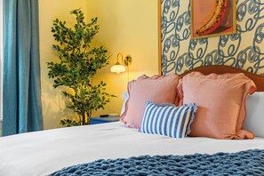 Ein Bett mit weißer Matratze, zwei Kissen, einer blauen Strickdecke, einer Wand mit blau-gelbem Muster, einer Zimmerpflanze und einer Wandlampe.
