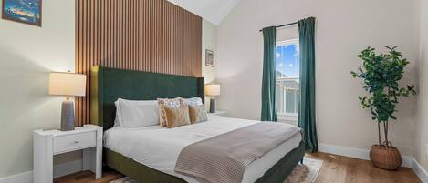 A bedroom with a bed, nightstands, a window with curtains, a plant, and a wall with vertical wooden slats.