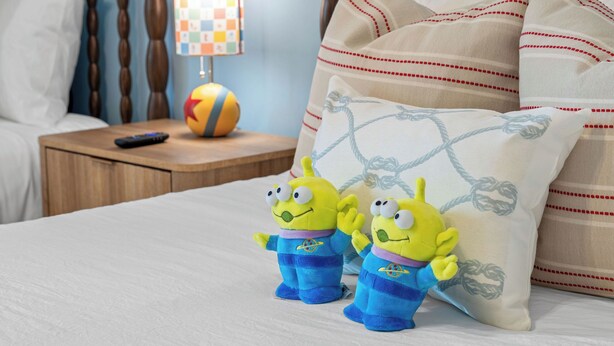 A bed with two plush toys, a nightstand with a remote control and a ball, and a pillow with a geometric pattern. - NEW Disney Lodge 4 min SDC+Hot tub • Sauna • Slide (Branson)