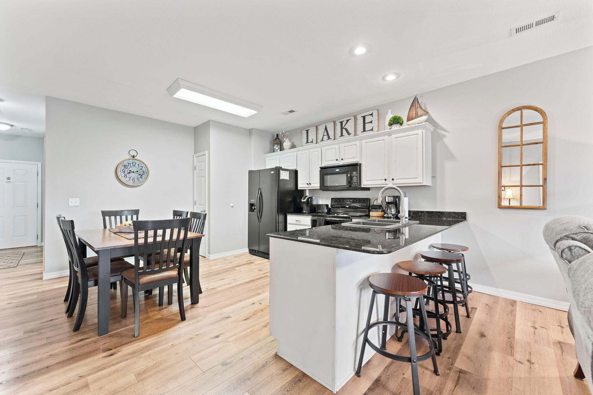 A modern kitchen with white cabinetry, a central island with bar stools, a dining area with a wooden table and chairs, and a clock on the wall.