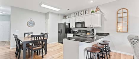 A modern kitchen with white cabinetry, a central island with bar stools, a dining area with a wooden table and chairs, and a clock on the wall.