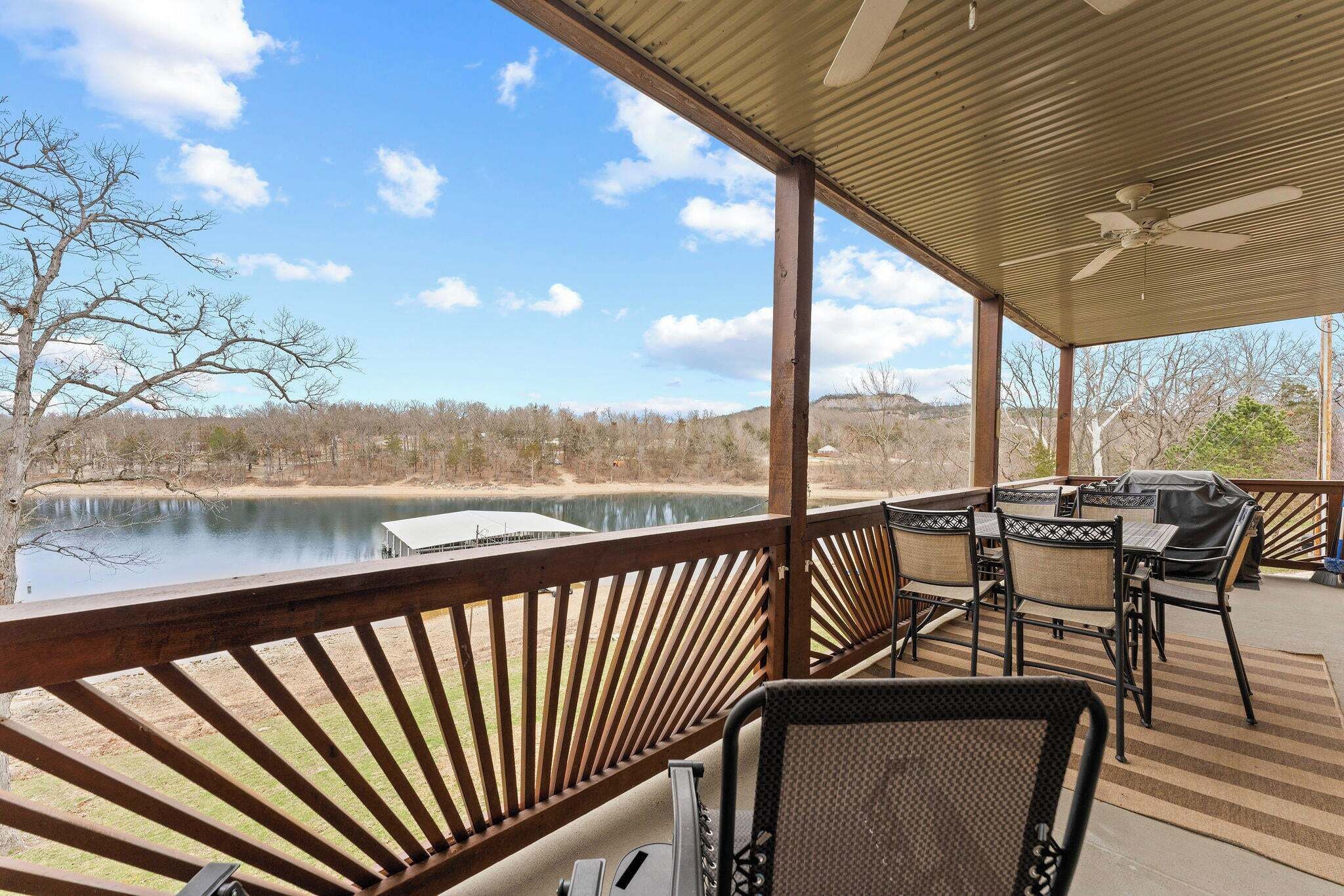 A covered outdoor deck with a view of a lake, a grill, and chairs.