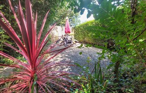 Uma trilha no jardim com uma planta vermelha em primeiro plano, uma área de pátio com mesa e cadeiras e um cercado de sebe ao fundo.