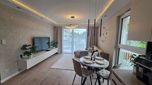 A modern living room with a flat-screen TV, a dining table set for four, and a balcony with a view.