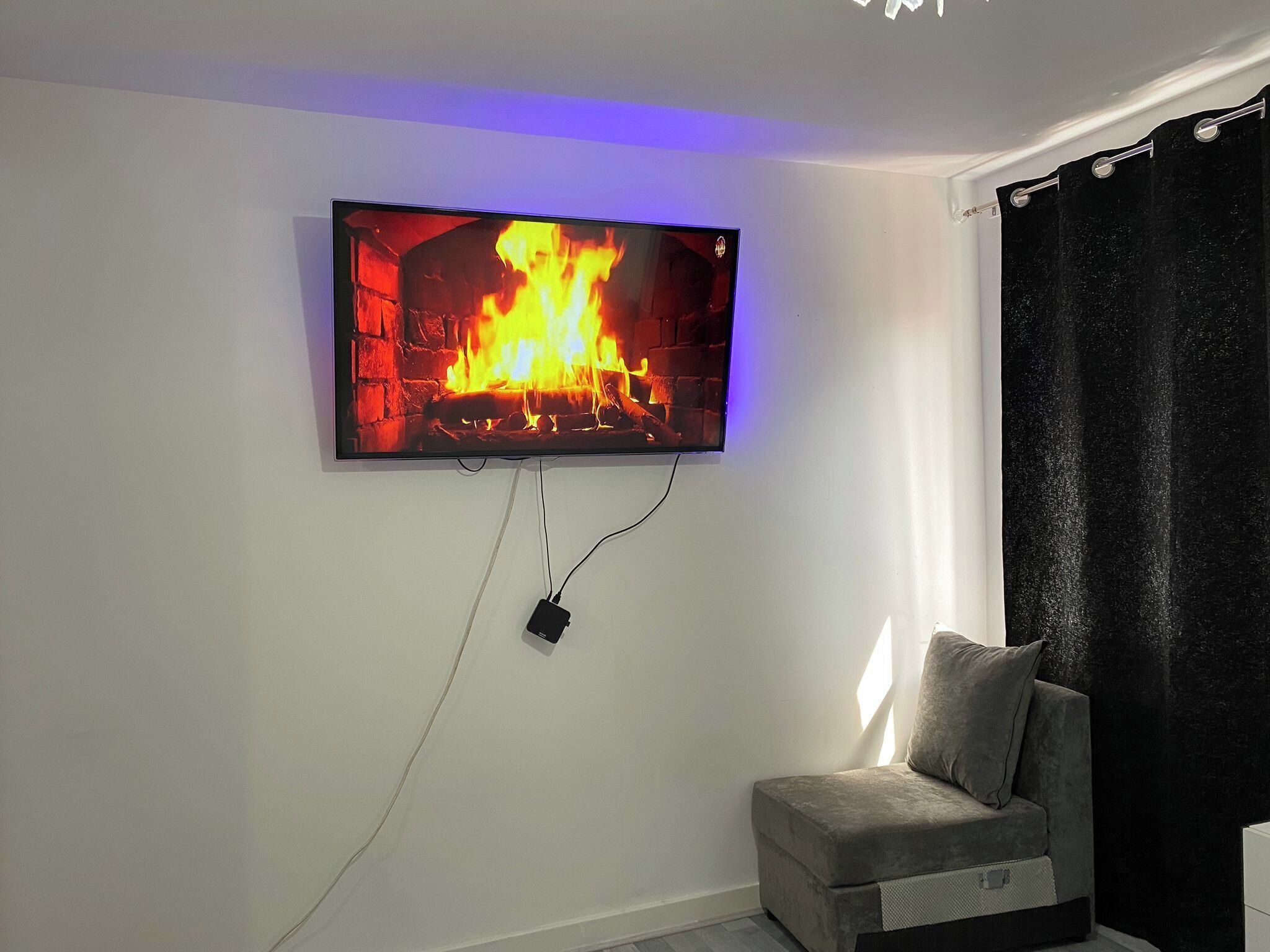 A television mounted on a wall displaying a fireplace image, a power outlet, and a grey armchair.