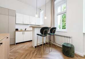 A modern kitchen with white cabinets, a wooden countertop, two black chairs, a green ottoman, and a window with a view of greenery. - Różana City Center Studio (Poznań)