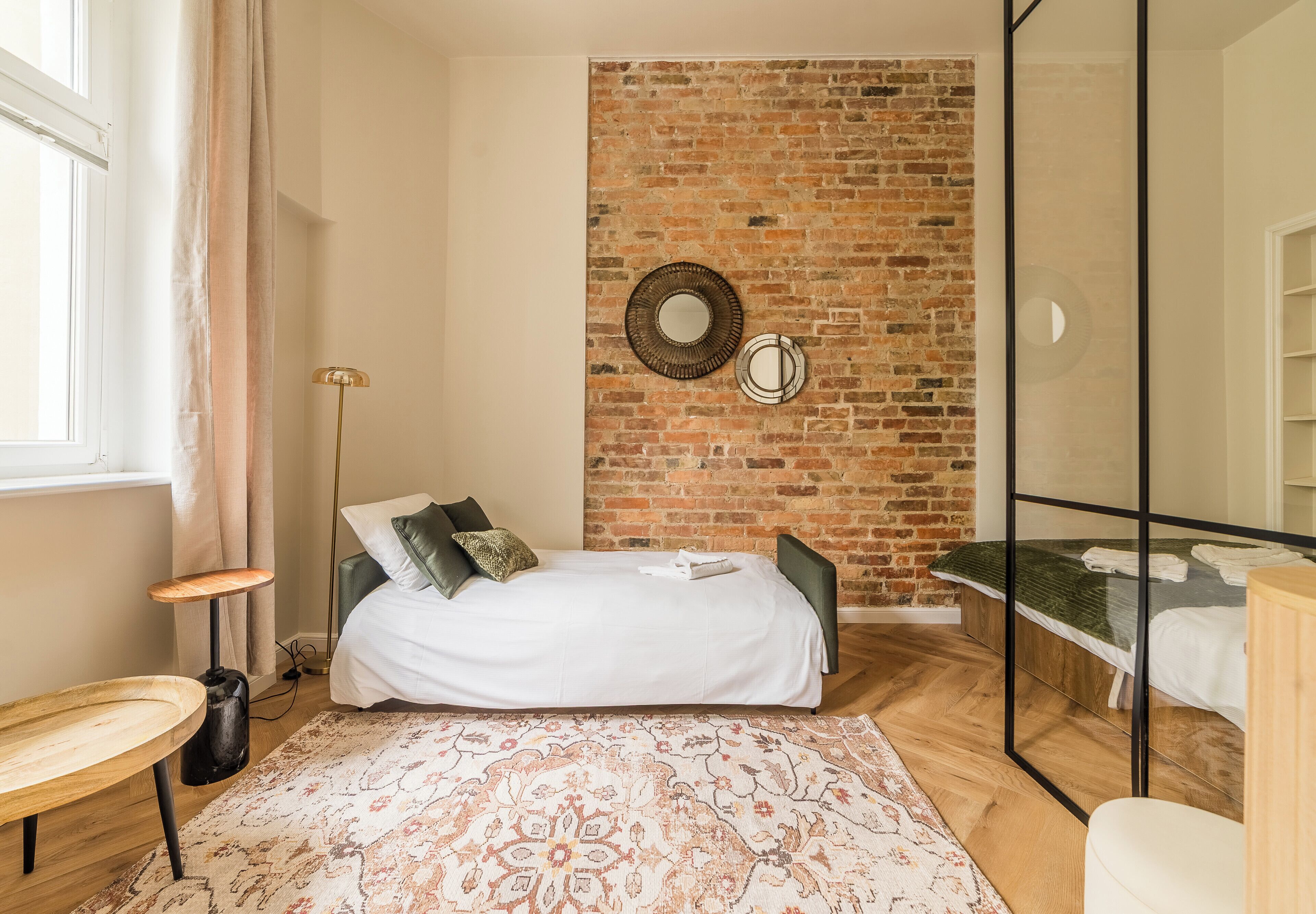 A bedroom with a brick wall, a bed with pillows, a bedside table, a floor lamp, and a patterned rug.