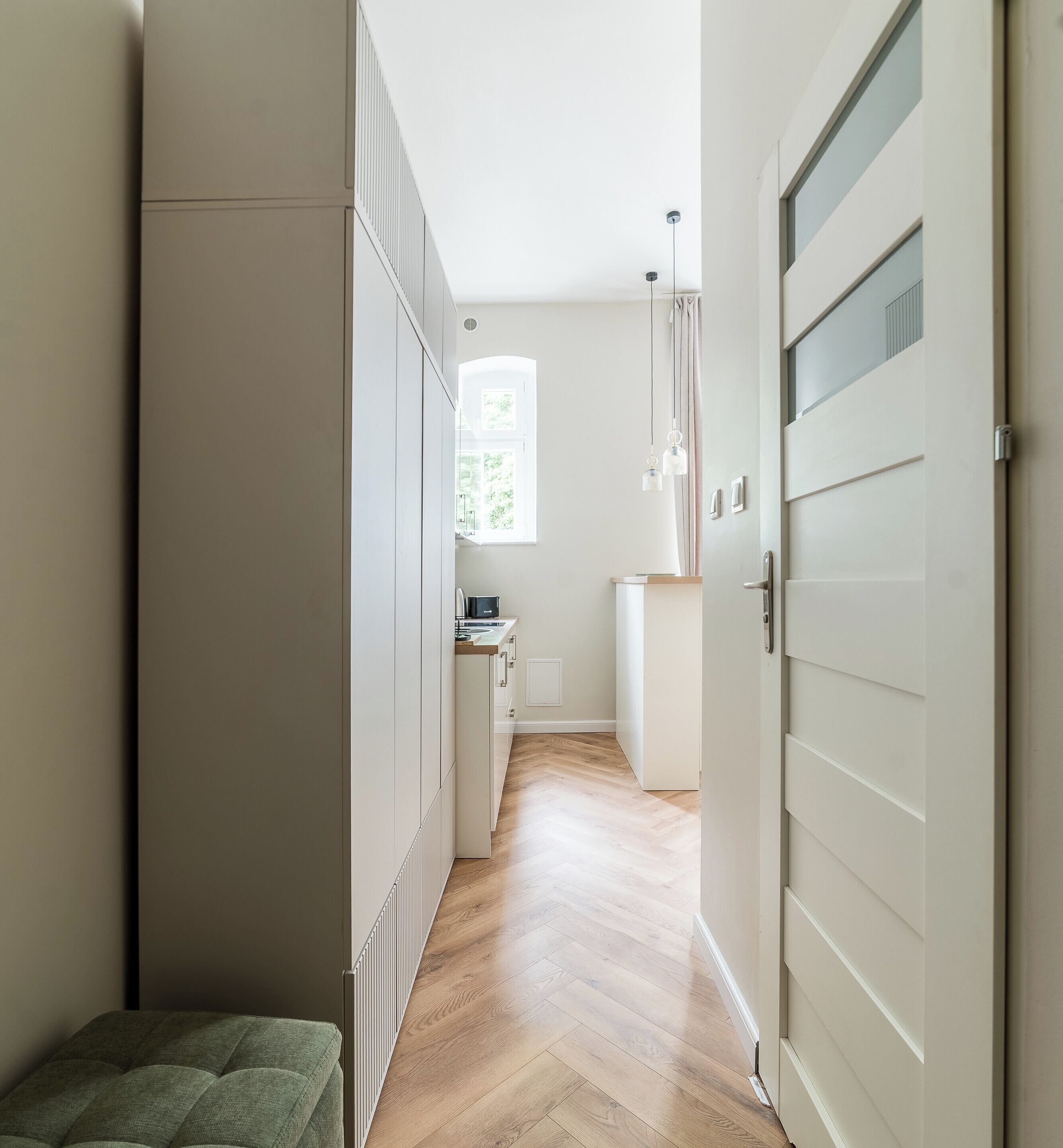 Un pasillo estrecho con un radiador, un banco y una puerta que da a una habitación con ventana.
