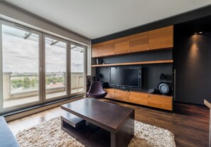 Een moderne woonkamer met een grote televisie, houten kasten, een bank en uitzicht door middel van kamerhoge ramen.