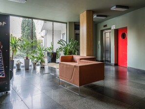 Un hall con un sofá marrón, plantas en macetas, una puerta roja y un ascensor.