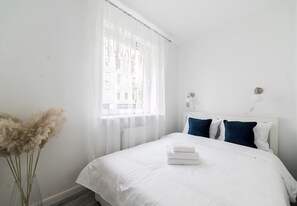 A bedroom with a bed, white bedding, a window with sheer curtains, and a vase with dried plants. - Sikorskiego Signature Apartment (Poznań)