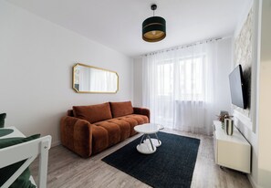 Un salon moderne avec un canapé marron, une table basse ronde blanche, un tapis noir, un meuble blanc sur lequel est posée une télévision à écran plat, et des rideaux légers.