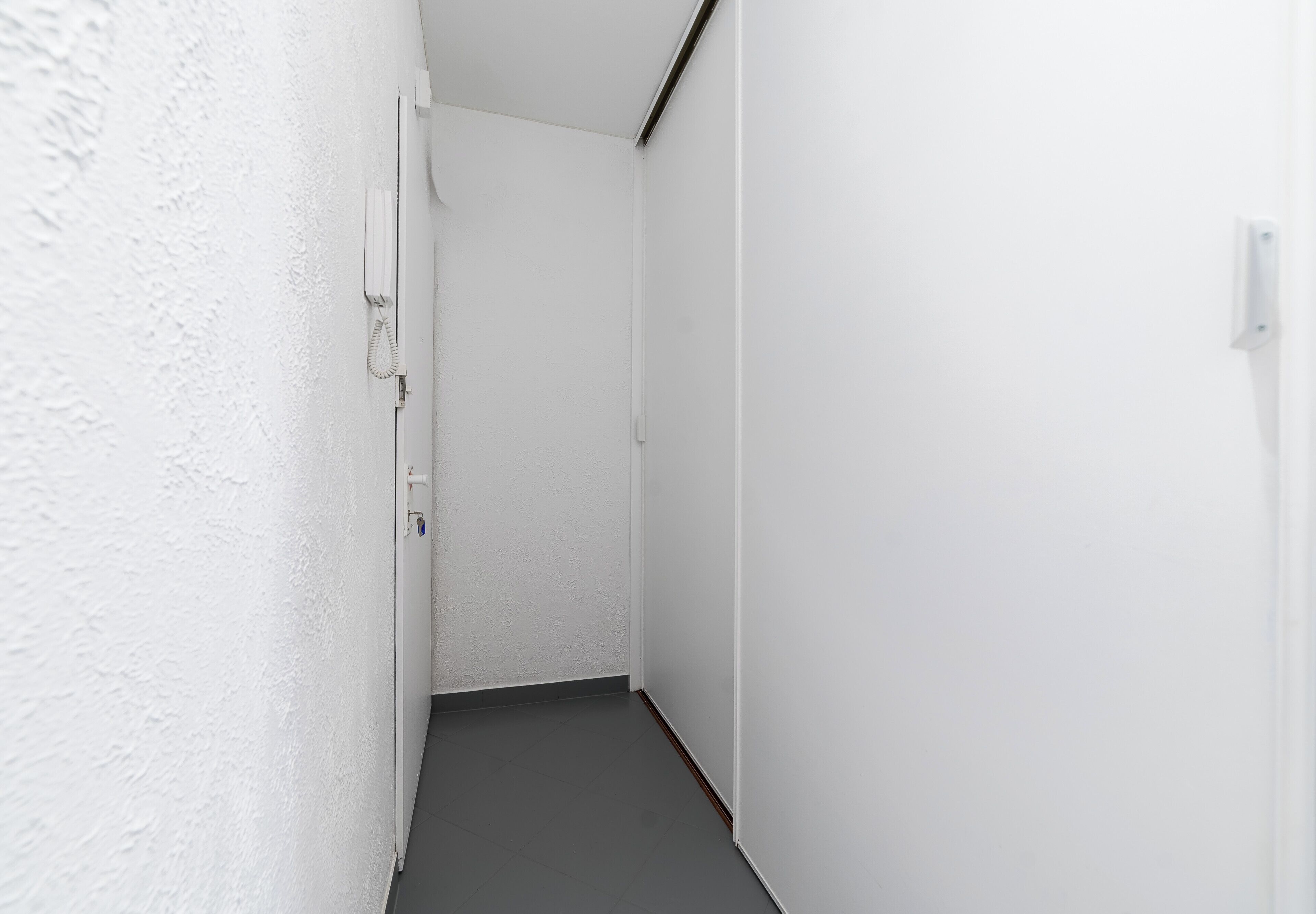 A narrow hallway with white walls and a dark floor, leading to a door at the end.