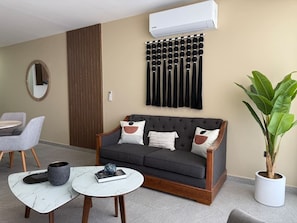 Ein modernes Wohnzimmer mit einem grauen Sofa, einem runden Couchtisch, einer Topfpflanze und einer Wand-Klimaanlage.