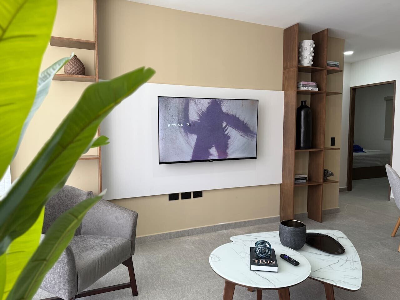 A modern living room with a flat-screen TV mounted on the wall, a grey armchair, a small coffee table with a remote control, and a bookshelf with books and decorative items.