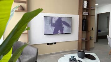 A modern living room with a flat-screen TV mounted on the wall, a grey armchair, a small coffee table with a remote control, and a bookshelf with books and decorative items.
