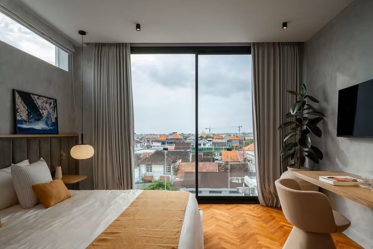 Ein modernes Hotelzimmer mit großem Fenster, einem Bett, einem Schreibtisch und einem Sessel.