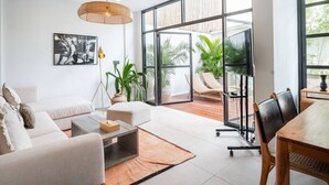A modern living room with a sofa, coffee table, and dining area. There is a large window providing natural light and a view of greenery outside. - Tribeca Bali (Bali)