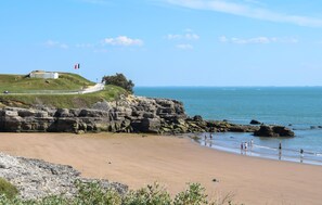 Un paysage côtier comprenant une plage de sable, des falaises rocheuses et une colline herbeuse sur laquelle se trouve une construction.
