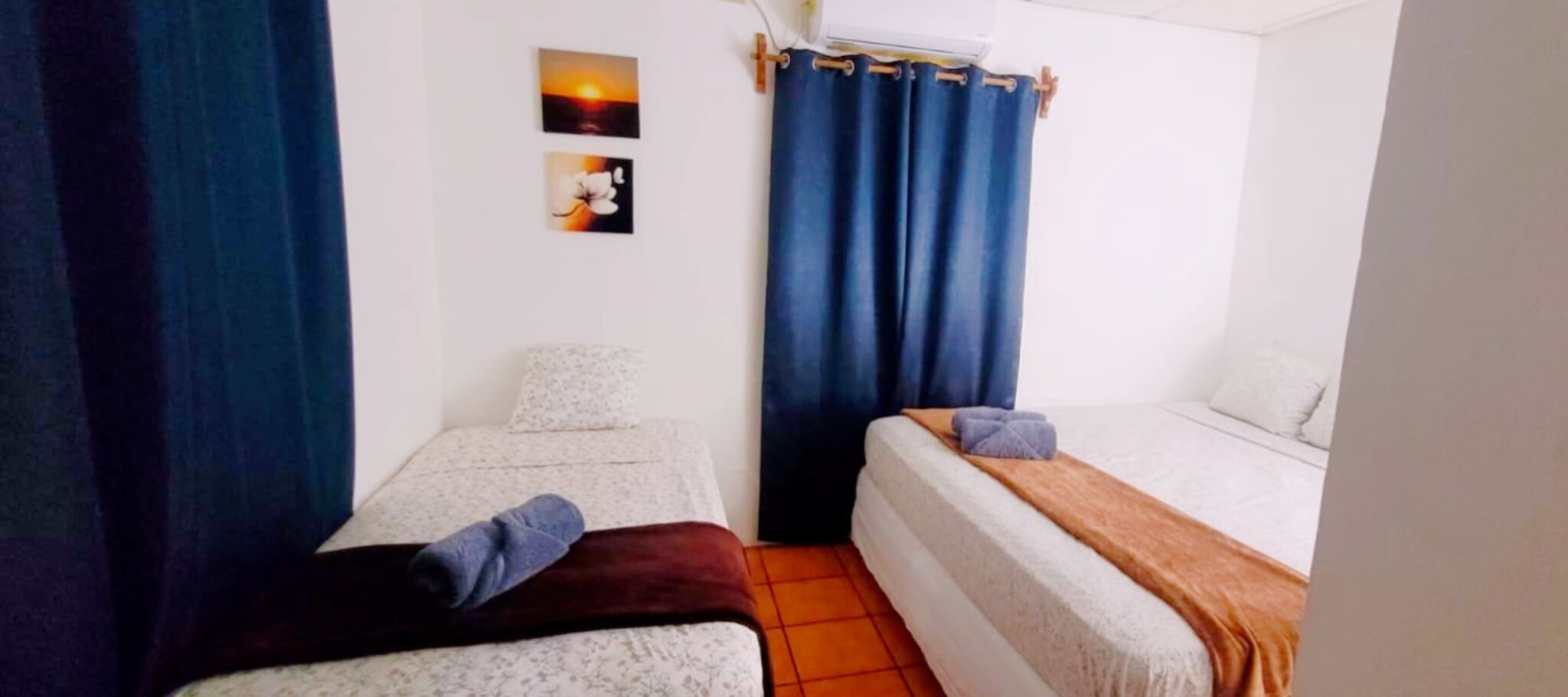 Een schone, kleine kamer met twee bedden, een raam met gordijnen en een wandairconditioner.