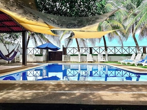 Piscina con sillas de descanso y una hamaca, rodeada de palmeras y una zona sombreada.