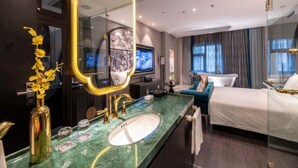 Habitación de hotel con un espejo grande, cama, televisor, baño con lavamanos y espejo, y ventana con cortinas.