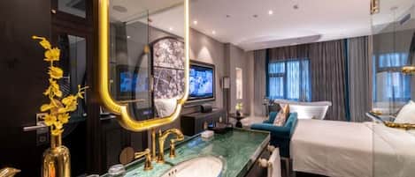 Habitación de hotel con un espejo grande, cama, televisor, baño con lavamanos y espejo, y ventana con cortinas.