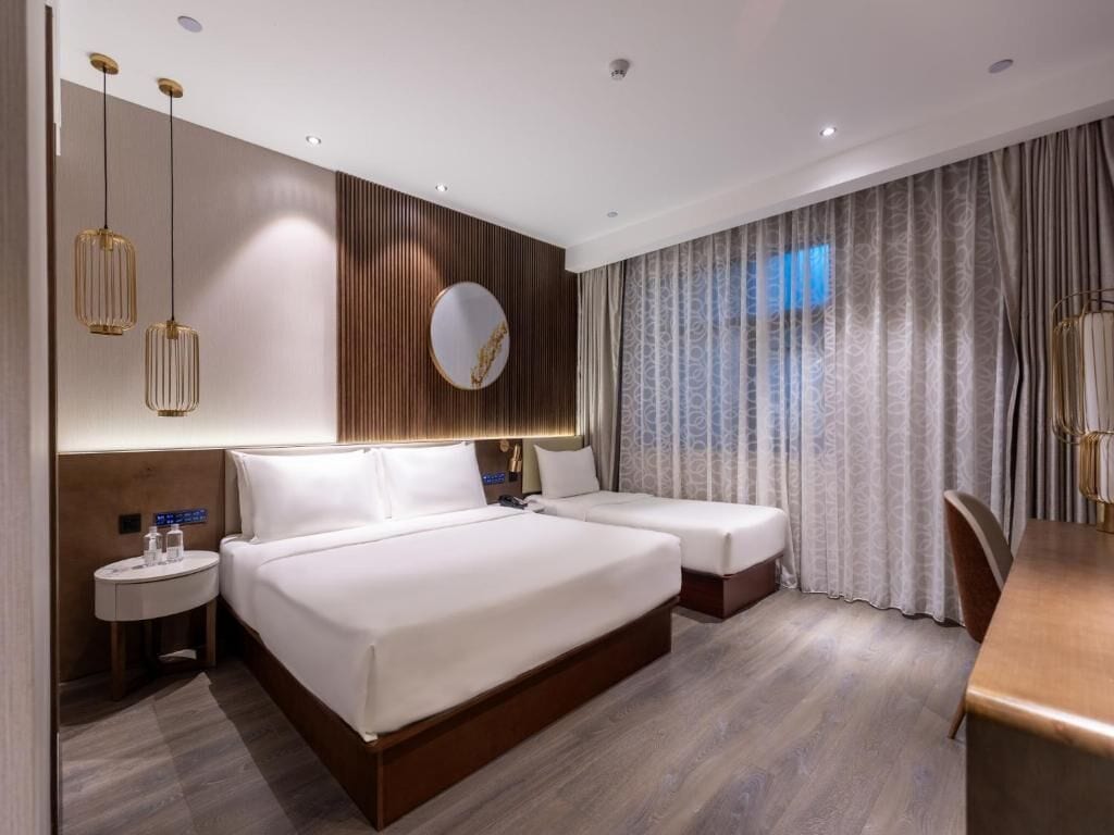 Une chambre d’hôtel moderne avec un grand lit, des tables de chevet, un miroir rond et une vue sur l’extérieur à travers des rideaux.