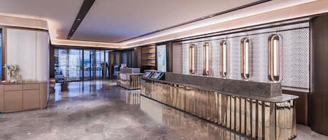 A modern hotel lobby with a reception desk, a seating area, and a view of the street through large windows.