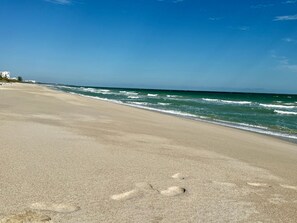 A beach with footprints leading to the water, a clear sky, and buildings in the distance. - Beachfront Island Escape- Heated Pool (Fort Pierce)