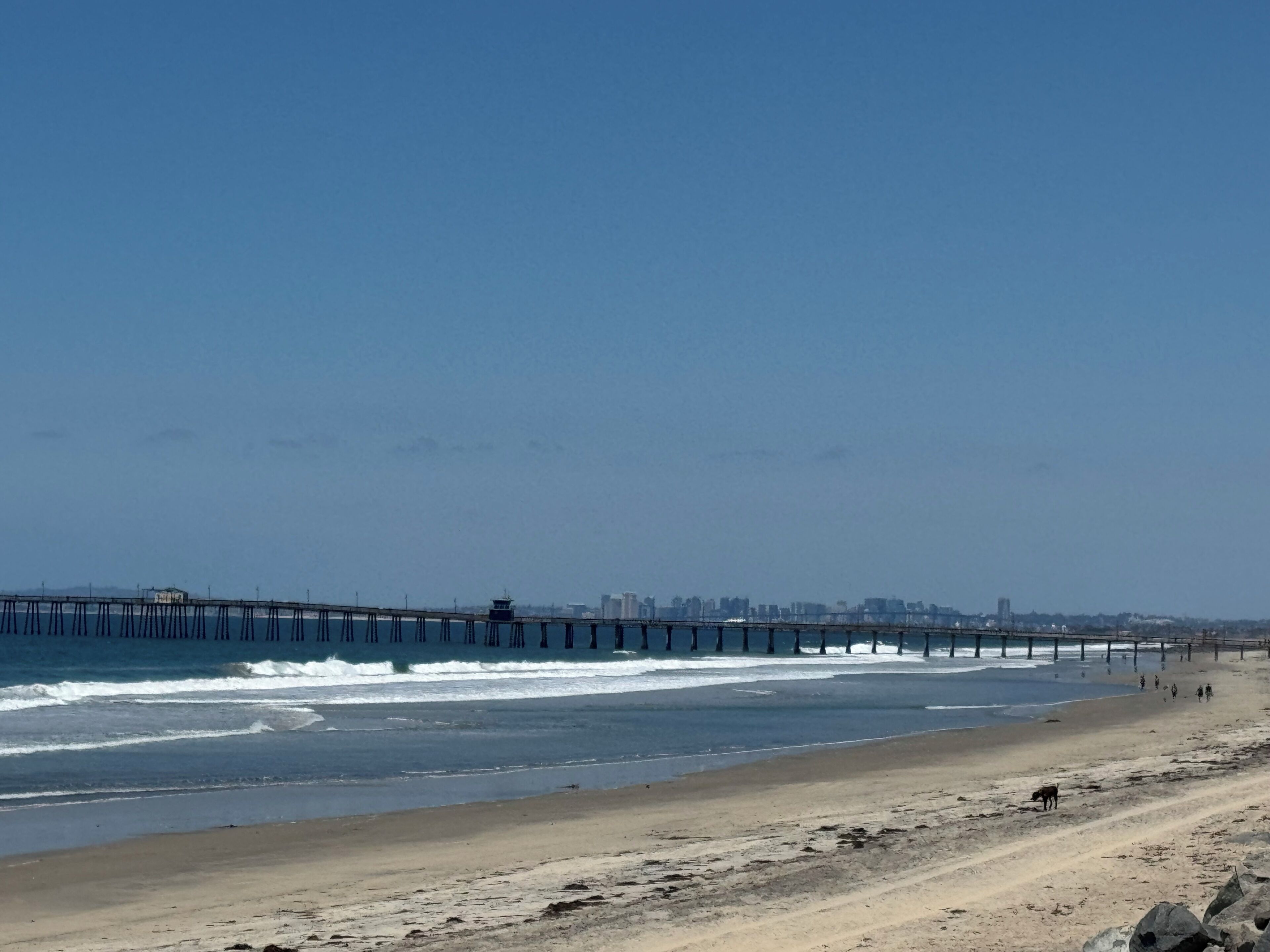Ein Strand mit einem Steg, der sich ins Meer erstreckt, einem sandigen Ufer und einer entfernten Stadtlandschaft.