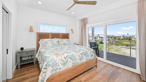 A bedroom with a bed, a nightstand, a ceiling fan, and a large window with a view of the outside. - Double D's by the Sea | New 2BR Coastal Retreat (Mexico Beach)