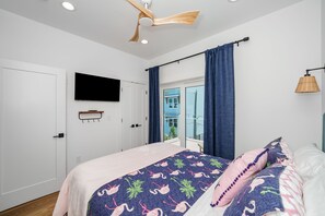A bedroom with a bed, a mounted TV, a ceiling fan, and a view of the outside. - Double D's by the Sea | New 2BR Coastal Retreat (Mexico Beach)