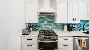 A kitchen with white cabinets, a gas stove, an oven, a coffee maker, and a blue tiled backsplash. - Double D's by the Sea | New 2BR Coastal Retreat (Mexico Beach)