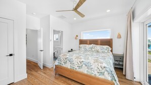 A bedroom with a wooden bed, a ceiling fan, a window with a view, and a door leading to another room. - Double D's by the Sea | New 2BR Coastal Retreat (Mexico Beach)