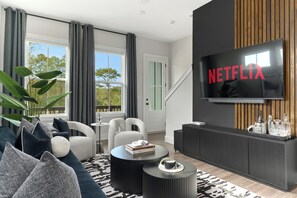 Ein modernes Wohnzimmer mit großem Flachbildfernseher an einer dunklen Wand, einer Couch mit gemusterten Kissen, einem Couchtisch und einer Topfpflanze.