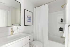 Une salle de bain moderne avec un lavabo, des toilettes et une baignoire, tous de couleur blanche. Un tableau abstrait encadré est accroché au-dessus du lavabo, et il y a un rideau de douche.
