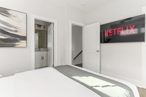 Ein Schlafzimmer mit einem Bett, einer Tür zu einem Badezimmer und einem an der Wand befestigten Fernseher mit dem Netflix-Logo.