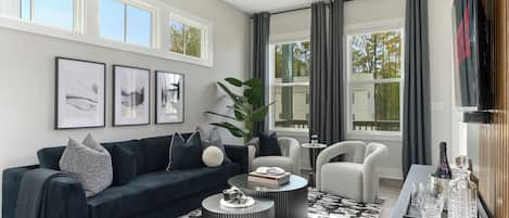 Una sala moderna con un sofá azul oscuro, dos mesitas redondas, una planta grande y un televisor sobre un mueble de madera.