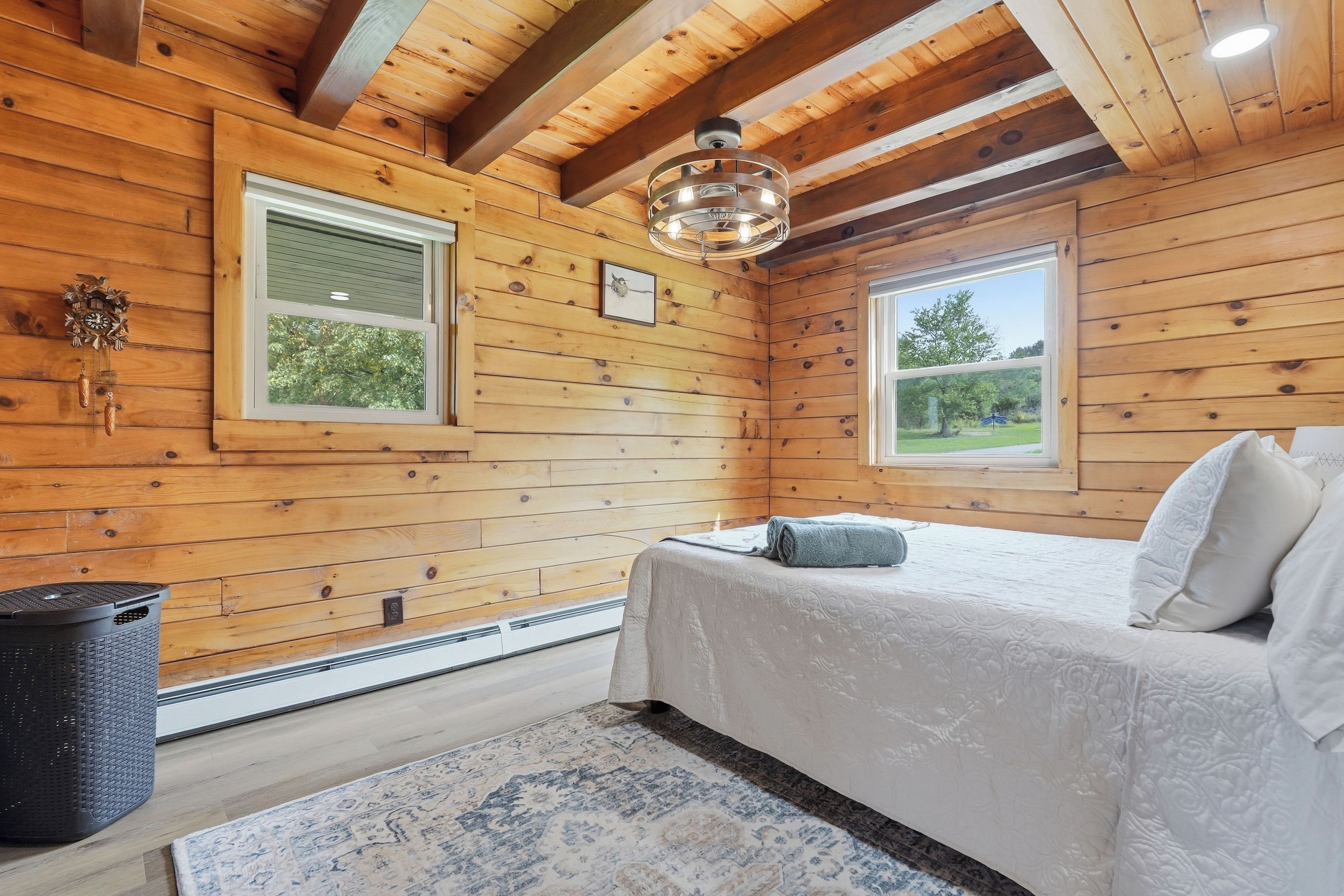 Un dormitorio de cabaña de madera con una cama, dos ventanas, una lámpara en el techo y un cesto de basura.