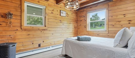 A wooden cabin bedroom with a bed, two windows, a ceiling light, and a trash bin.