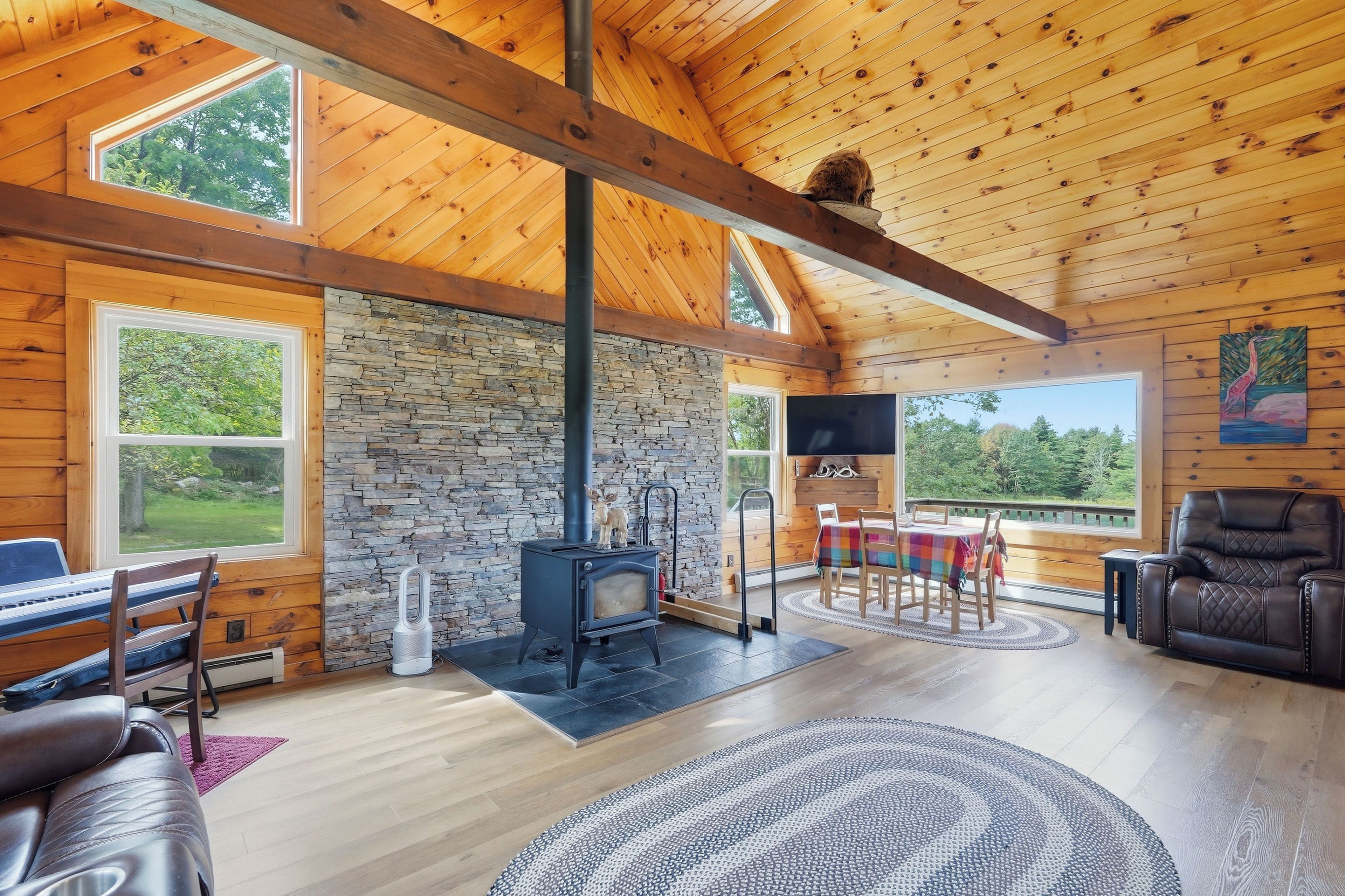 Amplia sala de estar con pared de piedra, vigas de madera en el techo, chimenea y grandes ventanales con vistas al exterior.