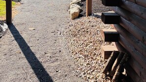 Ein überdachter Außenbereich mit einem Kiesweg, der zu einer Bank an einem See führt, umgeben von Bäumen und Holzbauten.
