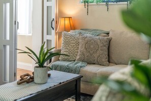 Ein gemütliches Wohnzimmer mit einer Couch, gemusterten Kissen, einer Lampe, einer Pflanze und einem kleinen Tisch.
